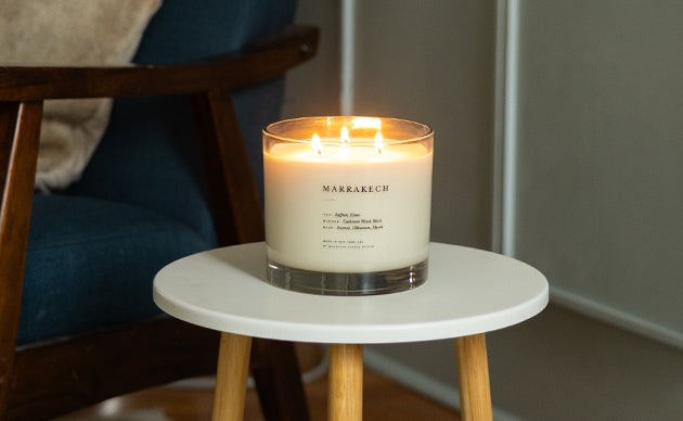 A lit three-wick candle in a clear glass jar sits on a small white side table in a cozy indoor setting with a blue chair in the background.
